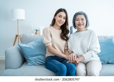 Portrait Of Asian Lovely Family, Young Daughter Hold Hand Older Mother. Attractive Girl And Senior Elder Mature Mom Sit On Sofa Enjoy Activity At Home Together In Living Room, Smile And Look At Camera