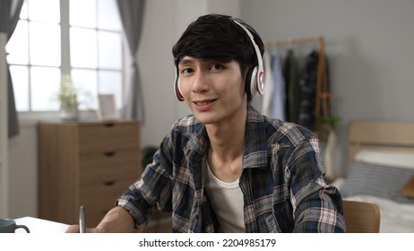 Portrait Of Asian Male Adult Student Learning From Home Is Turning To Look At Camera With A Smile While Writing Homework In The Bedroom During Lockdown