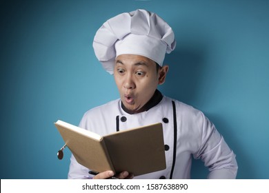 Portrait Of Asian Male Chef Reading Book Of Recipes, Finding Secret Recipe, Against Blue Background