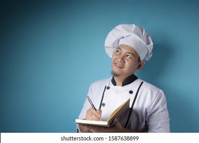 Portrait Of Asian Male Chef Reading Book Of Recipes, Finding Secret Recipe, Against Blue Background