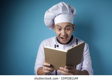 Portrait Of Asian Male Chef Reading Book Of Recipes, Finding Secret Recipe, Against Blue Background