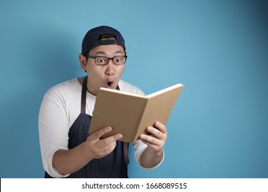 Portrait Of Asian Male Chef Reading Book Of Recipes, Finding Secret Recipe, Against Blue Background