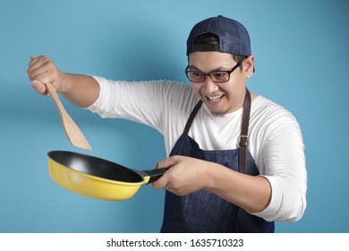 Portrait Of Asian Male Chef Smiling Happy While Holding Kitchen Tools, Chef Holding Spatula And Pan, Ready To Cook, Against Blue Background