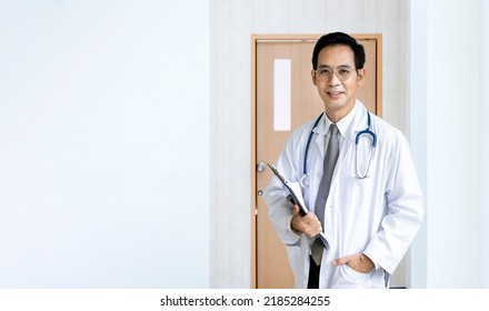 Portrait Of Asian Male Doctor At The Hospital Ward In Front Of Patient Room Holding Clipboard With Medical Record For Better Nursing Care Plan With Copy Space