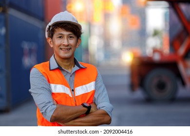 Portrait Asian male industrial engineer in white helmet, orange safety vest standing smile and arms crossed look at camera. Supervisor or large warehouse management specialists in container terminal. - Powered by Shutterstock