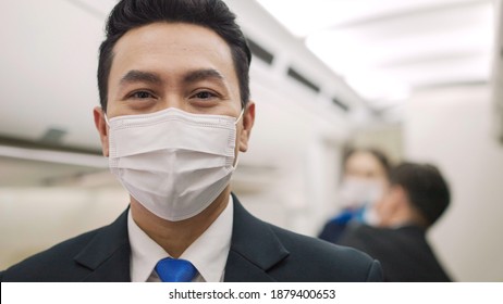 Portrait Of Asian Male Steward Wearing Medical Face Mask To Prevent Coronavirus On Airplane Cabin. Airline Business Responsibility Service And Support Travel During Virus Epidemic