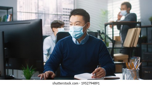 Portrait Of Asian Male Worker Sitting At Desk In Cabinet Typing On Computer And Writing In Planner, Work In Coronavirus Quarantine. Colleagues On Background. Man In Medical Mask Work In Company Office