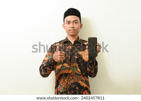 portrait of an Asian man in batik shirt and cap holding and displaying a smartphone screen. Indonesian Muslim man showing cellphone screen and showing thumbs up on isolated white background