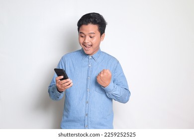Portrait of Asian man in blue shirt raising his fist, celebrating success, winning game on his mobile phone. Isolated image on white background - Shutterstock ID 2259252525