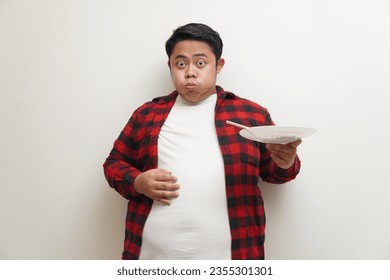 Portrait of Asian man in red plaid shirt feeling full after eating while holding his stomach. Isolated on white background