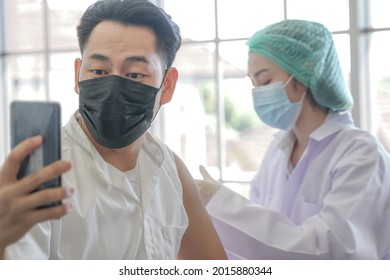 Portrait Of Asian Man Selfie By Phone At While Getting Covid Vaccine In Clinic Or Hospital, With Hand Nurse Injecting Vaccine To Get Immunity For Protect Virus. People Wearing Protective Mask.