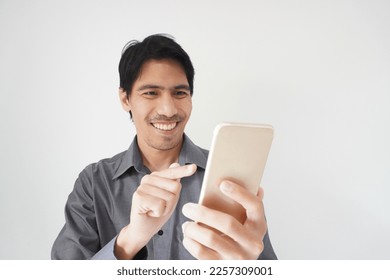 Portrait of an Asian man smiling, holding a smartphone in his hand, cheerful with the text message. Isolated on grey background. Studio concept of a male model with smart internet communication. - Shutterstock ID 2257309001