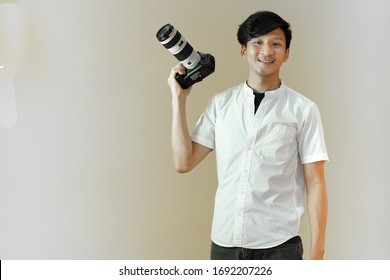 Portrait of asian man in white shirt holding digital camera with smile. Handsome male photographer standing isolated on plain background copy space. Photography business concept. - Powered by Shutterstock