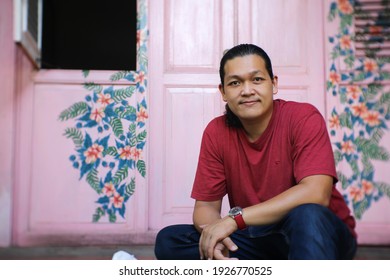 Portrait Of Asian Mature Business Man With Black Long Hair And Casual Clothes Sit In Front Of Pink Vintage Wooden Door. Modern Internet Billionaire Who Working From Home