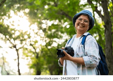 Portrait of Asian mature woman holding a camera and backpack behind her back, an Asia active senior woman enjoying nature in park. Standing on a trail in a forest outdoors. Enjoying active travel trip - Powered by Shutterstock