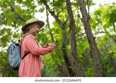 Portrait of Asian mature woman holding smartphone and backpack behind back, an Asia active senior woman enjoying nature in park. Standing on a trail in a forest outdoors. Enjoying active travel trip - Powered by Shutterstock