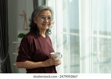 Portrait of Asian mature woman standing near curtain with sunlight relaxing with cup of coffee in living room, Senior woman with eyeglasses smiling and looking at camera at home., copy space - Powered by Shutterstock