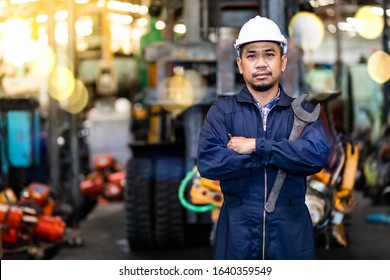 Portrait Of Asian Mechanic Fold Over Holding A Wrench And Smiling At Truck And Forklift Garage. Industrial Mechanic Engineer In Hard Hat