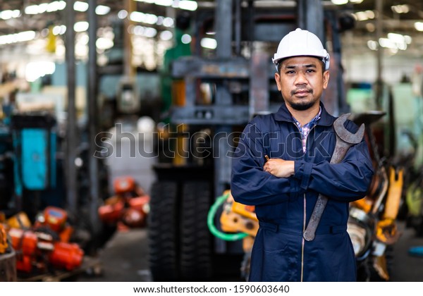Portrait of Asian mechanic Fold over holding a wrench\
and smiling at truck and forklift garage. Industrial mechanic\
Engineer in Hard Hat
