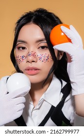 Portrait of asian model with glitter visage holding balls and looking at camera isolated on orange