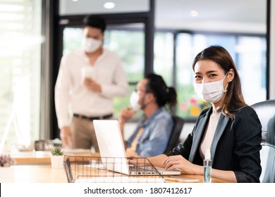 Portrait of asian office employee businesswoman wear protective face mask work in new normal office with interracial colleague in background as social distance practice prevent coronavirus COVID-19.