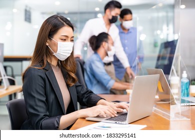 Portrait of asian office employee businesswoman wear protective face mask work in new normal office with interracial team in background as social distance practice prevent coronavirus COVID-19.