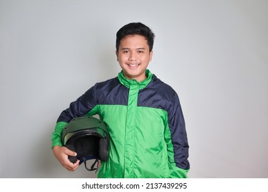 Portrait Of Asian Online Taxi Driver Wearing Green Standing Against Gray Background, Smiling And Looking At Camera, Holding His Helmet On The Side