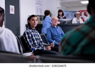Portrait Of Asian Patient In Hospital Reception Lobby, Sitting In Waiting Area To Attend Doctor Appointment In Private Hospital. Young Adult In Medical Clinic With Diverse Patients.