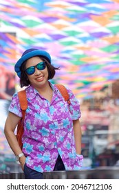 Portrait Of Asian Plus Size LGBT Tourist In Hawaiian Shirt With Sunglasses Smiling On Walking Street With Colorful Decorative Fabric Hanging On The Air During Summer Holiday