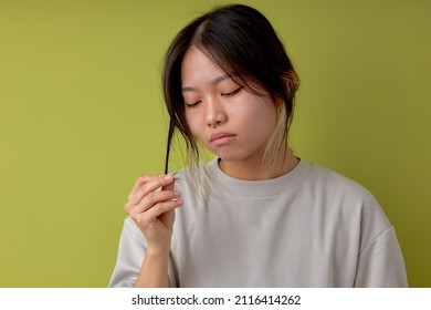 Portrait Of Asian Sad Woman Touching Hair, Stand With Thoughtful Facial Expression, Isolated Over Green Studio Background. Chinese Lady In Casual Beige T-shirt Want To Cut Hair. Natural Beauty