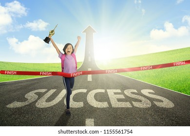 Portrait of Asian schoolgirl carrying a trophy while crossing the finish line on the road with success word and upward arrow - Powered by Shutterstock