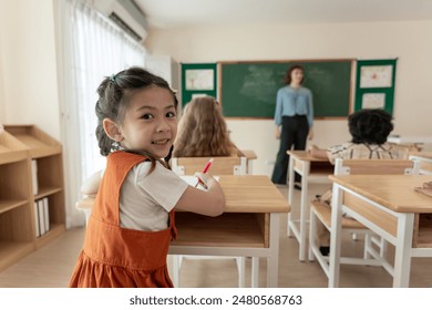 Portrait of Asian schoolgirl learning in classroom at elementary school. Young adorable kid student sitting on study table and listen educate subject from teacher at kindergarten then look at camera. - Powered by Shutterstock