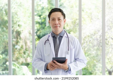 Portrait Of Asian Senior Doctor Standing In Medical Office While Using Tablet.