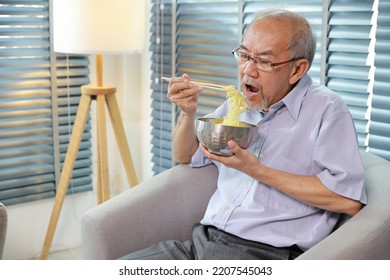 Portrait Of Asian Senior Man With Beard Sitting On Sofa Eating Tasty And Instant Noodle For Lunch In Living Room With Window Blinds. Unhealthy And Cheap Food Lifestyle Concept