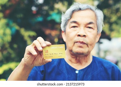 Portrait Of An Asian Senior Man, More 60 Years Old, Holding Golden Credit Card And Showing On Hand And Looking At Camera Feeling Enjoy. Elderly Financial Lifestyle Concept With Copy Space In Left Side