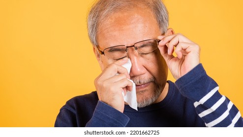 Portrait Asian Senior Old Man Sad Wiping Away His Tears Studio Shot Isolated On Yellow Background, Elder Man Crying Raise Glasses With Tissue Wipe Red Eyes, Sadness Depressed Lonely