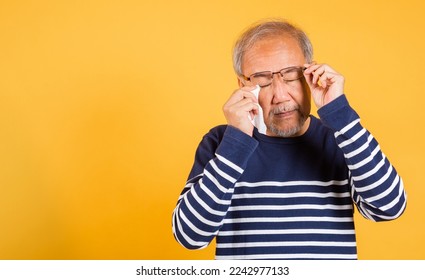 Portrait Asian senior old man sad wiping away his tears studio shot isolated on yellow background, Elder man crying raise glasses with tissue wipe red eyes, Sadness depressed lonely - Powered by Shutterstock