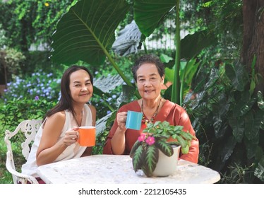 Portrait Of Asian Senior Woman And Her Daughter  Sitting Together At White Table In Beautiful Garden, Drinking Coffee From Blue And Orange Cup, Smiling And Looking At Camera.