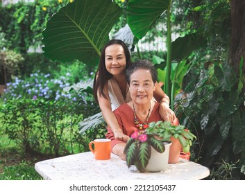 Portrait Of Asian Senior Woman Sitting With Her Daughter  At White Table In Beautiful Garden, Daughter Hugging Her Mother From The Back,  Smiling And Looking At Camera.