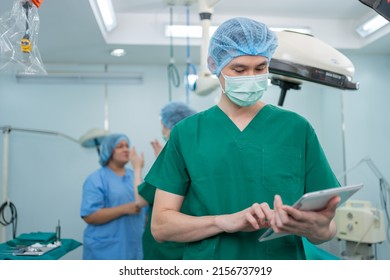 Portrait Of Asian Surgeon With Medical Mask Standing And Holding A Tablet In Operation Theater At A Hospital. Team Of Professional Surgeons. Healthcare, Emergency Medical Service Concept