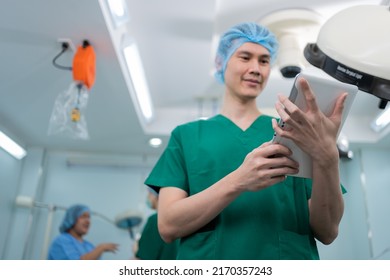 Portrait Of Asian Surgeon With Medical Mask Standing And Holding A Tablet In Operation Theater At A Hospital. Team Of Professional Surgeons. Healthcare, Emergency Medical Service Concept