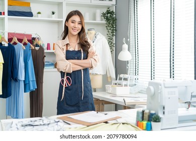 Portrait of Asian tailor woman working on clothes in tailoring atelier. Attractive beautiful young female fashion designer smile and look at camera after success in make new handmade suit in workshop.