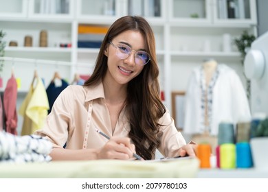 Portrait Of Asian Tailor Woman Working On Clothes In Tailoring Atelier. Attractive Female Fashion Designer Dressmaker Design Drawing And Sketching Out Ideas For New Fashion Collection In Workshop Room