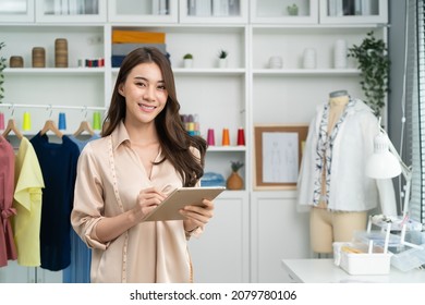 Portrait of Asian tailor woman working on clothes in tailoring atelier. Attractive beautiful young female fashion designer smile and look at camera after success in make new handmade suit in workshop. - Powered by Shutterstock