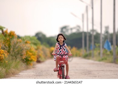 Portrait Of An Asian Thai Kid Girl Aged 4 To 6 Sharing A Red Bike In The Evening. She Enjoys Riding Bikes On The Road With A Happy And Bright Smile On Her Face. Cycling Is A Fun Exercise.