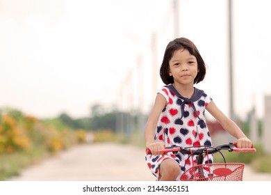 Portrait Of An Asian Thai Kid Girl Aged 4 To 6 Sharing A Red Bike In The Evening. She Enjoys Riding Bikes On The Road With A Happy And Bright Smile On Her Face. Cycling Is A Fun Exercise.