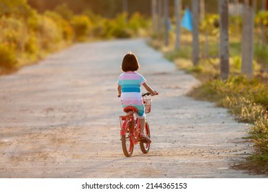 Portrait Of An Asian Thai Kid Girl Aged 4 To 6 Sharing A Red Bike In The Evening. She Enjoys Riding Bikes On The Road With A Happy And Bright Smile On Her Face. Cycling Is A Fun Exercise.