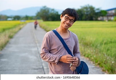 Portrait Of An Asian Thai Man Looking Humble And Relaxed Surrounded By Rice Field And In The Background There Is An Unknown Person Riding A Bicycle