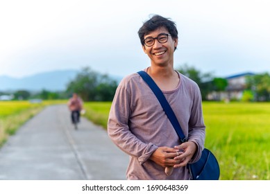 Portrait Of An Asian Thai Man Looking Humble And Relaxed Surrounded By Rice Field And In The Background There Is An Unknown Person Riding A Bicycle