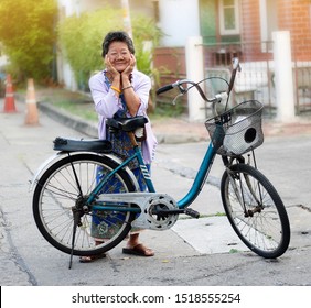 Portrait Asian Thai Senior Woman Smiling And Old Bicycle On Street Road. Happy Older Female Standing With Chin Resting On Hand Over Bike. Healthy Grandma And Sunset Blurred Housing Estate Background.